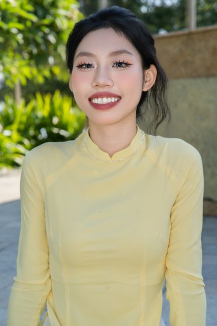 A cheerful young woman wearing a yellow ao dai poses outdoors in sunlight.