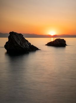 Peaceful ocean sunset view with rocks silhouetted against orange sky, calm waters.