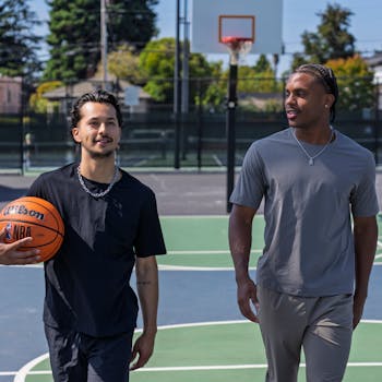 Two men walking on an outdoor basketball court, holding a basketball on a sunny day.