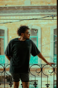Young man standing on balcony with urban backdrop, captured at sunset for a moody ambiance.