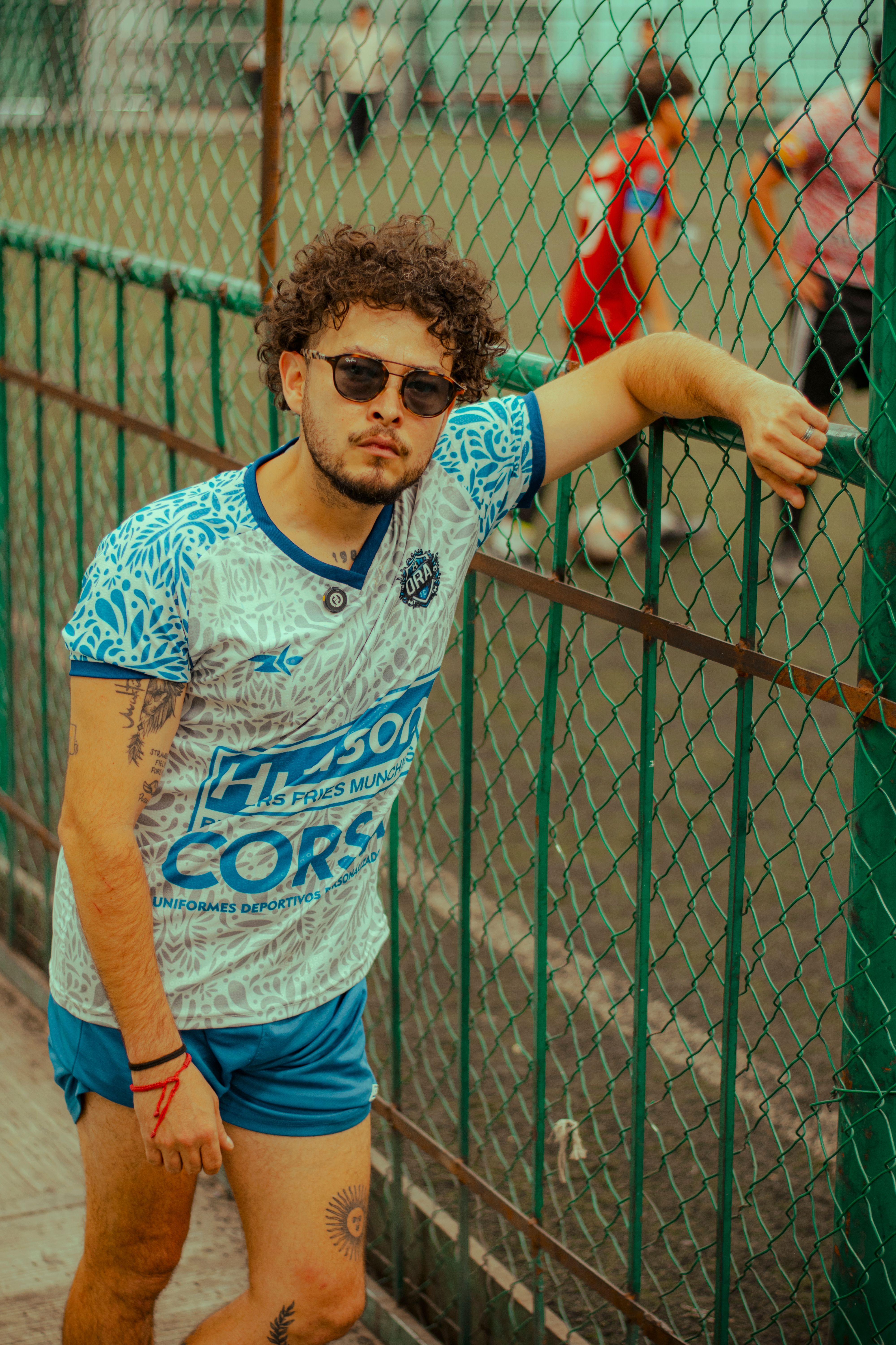 Stylish man in sportswear with tattoo leaning on a fence by a soccer field.