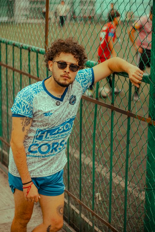 Free Stylish man in sportswear with tattoo leaning on a fence by a soccer field. Stock Photo