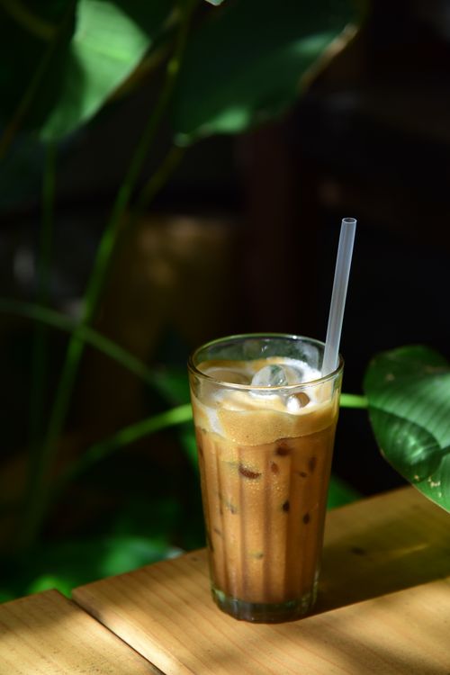 Free Chilled iced coffee with straw in bright café, surrounded by lush green leaves. Stock Photo