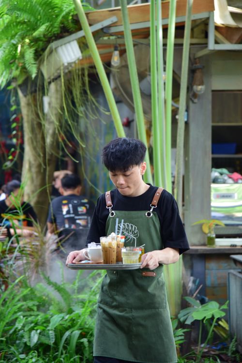 Free Barista in green apron serves drinks in a lush, outdoor café setting, capturing the essence of café culture. Stock Photo