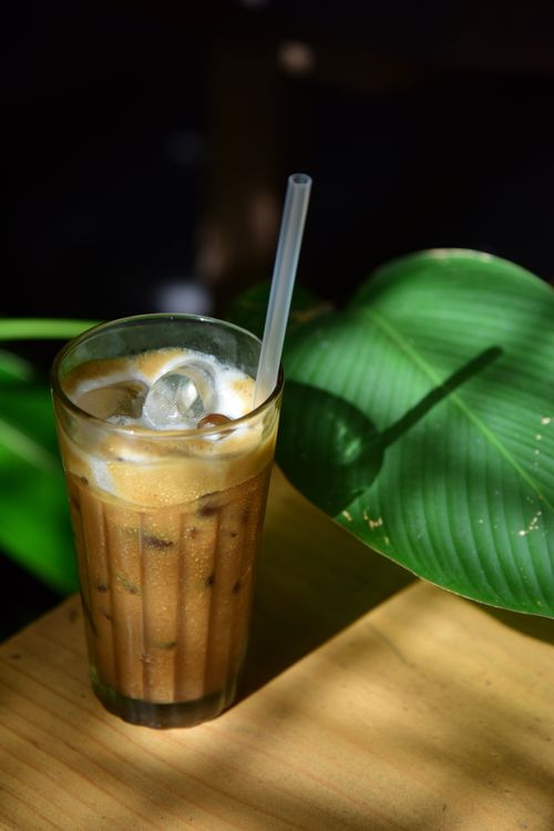 Free Tall glass of iced coffee with ice cubes beside a large green tropical leaf on a wooden table. Stock Photo