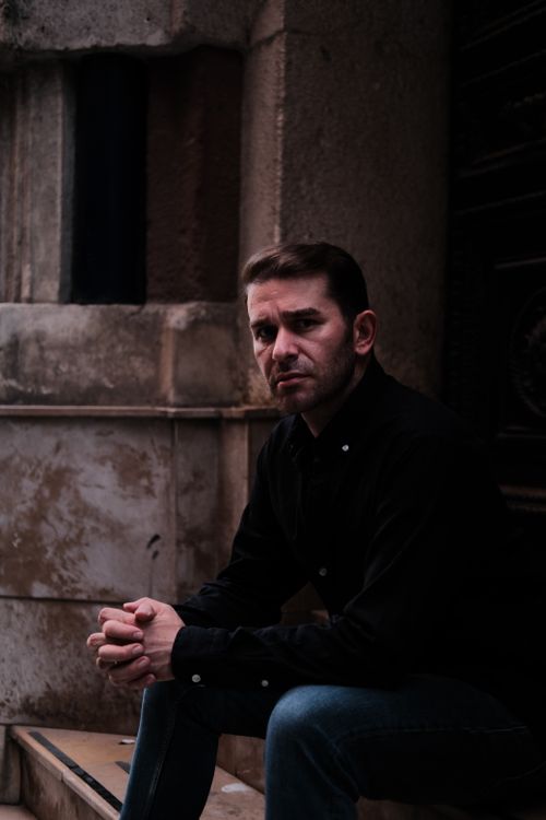 Free A thoughtful man sits against a stone wall in low light, wearing a black shirt and jeans. Stock Photo