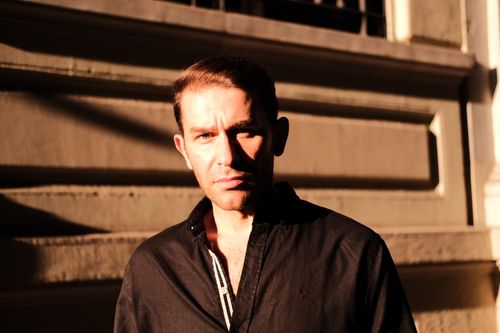 Free A man standing in sunlight against an urban building backdrop, casting long shadows. Stock Photo