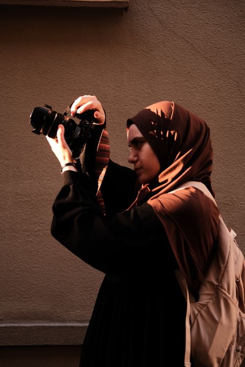 Free Side profile of a young woman in a hijab taking a photo. Warm lighting highlights her intent focus. Stock Photo