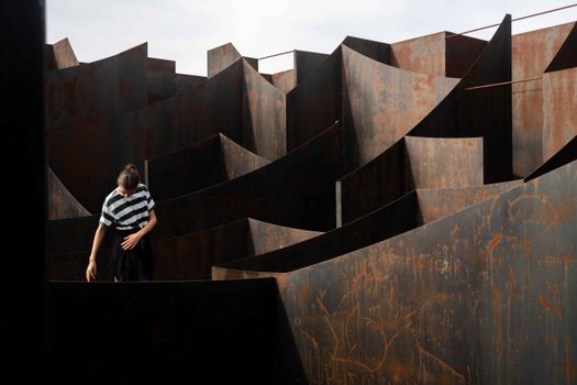 Person explores rusted metal labyrinth under tranquil daylight in Genk.