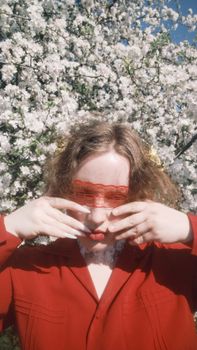 A woman in red with lace covering her eyes stands before blooming cherry blossoms.