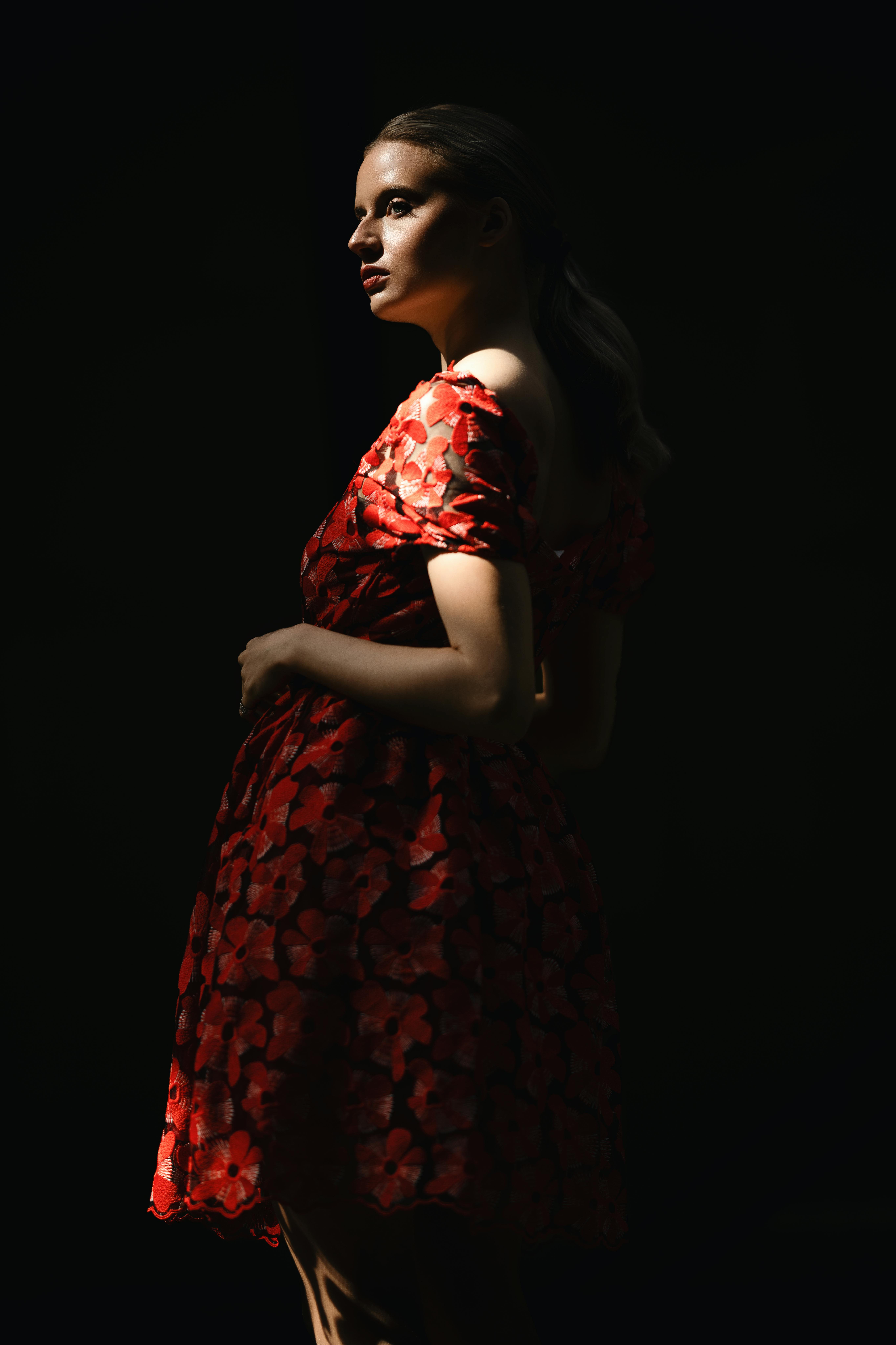A stylish woman in a red floral dress posing against a dark backdrop with dramatic lighting.