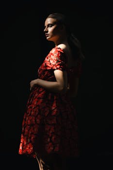 A stylish woman in a red floral dress posing against a dark backdrop with dramatic lighting.