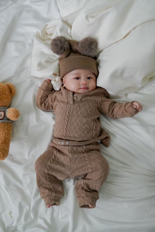 Free A cute baby lying on a white bed wearing a brown outfit and hat with pom-poms. Stock Photo