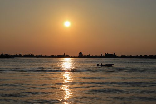 Free A tranquil sunset scene over a peaceful lake with a lone boat silhouetted against the glowing sky. Stock Photo