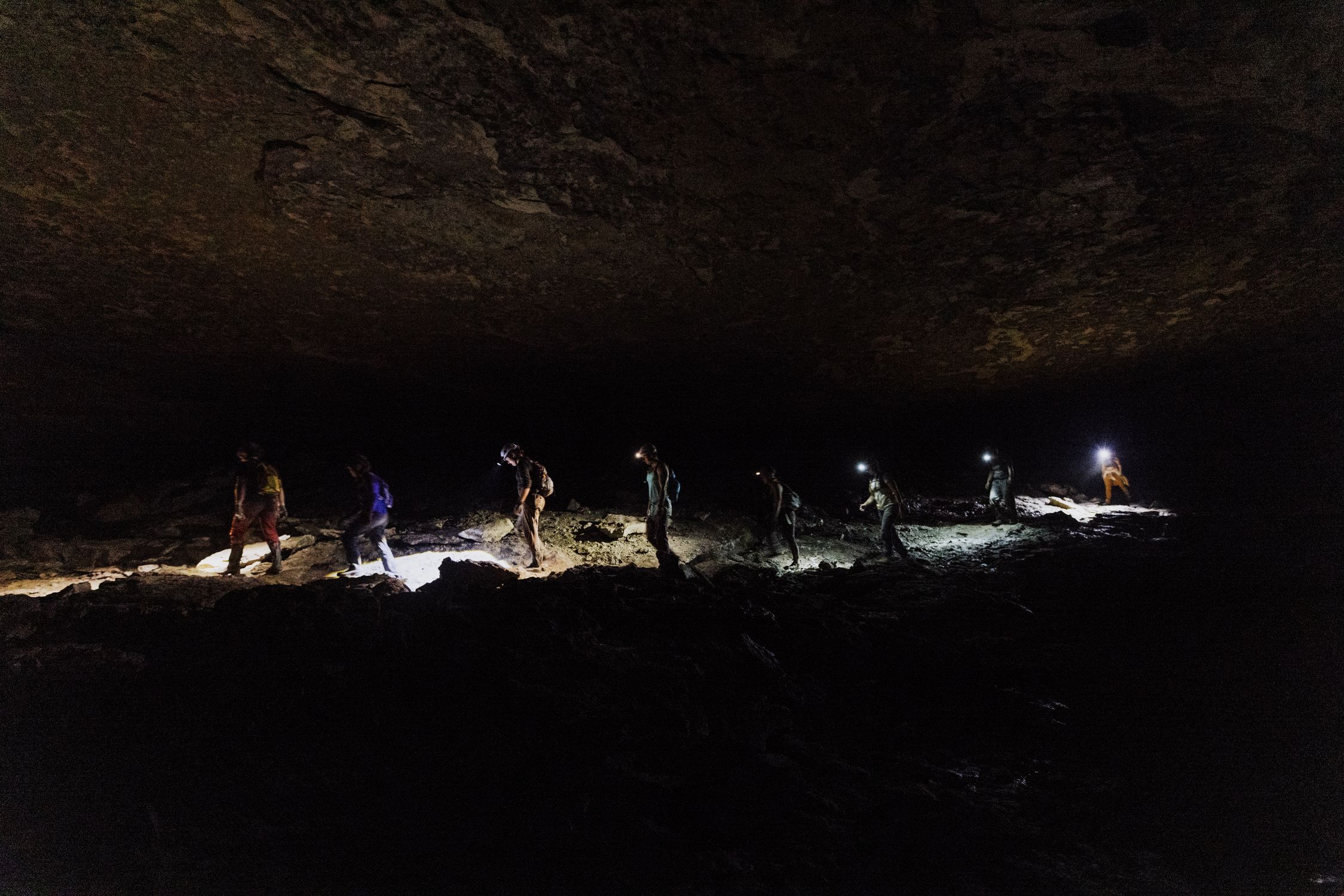 Adventurers explore a rugged dark cave in low light with headlamps and gear.