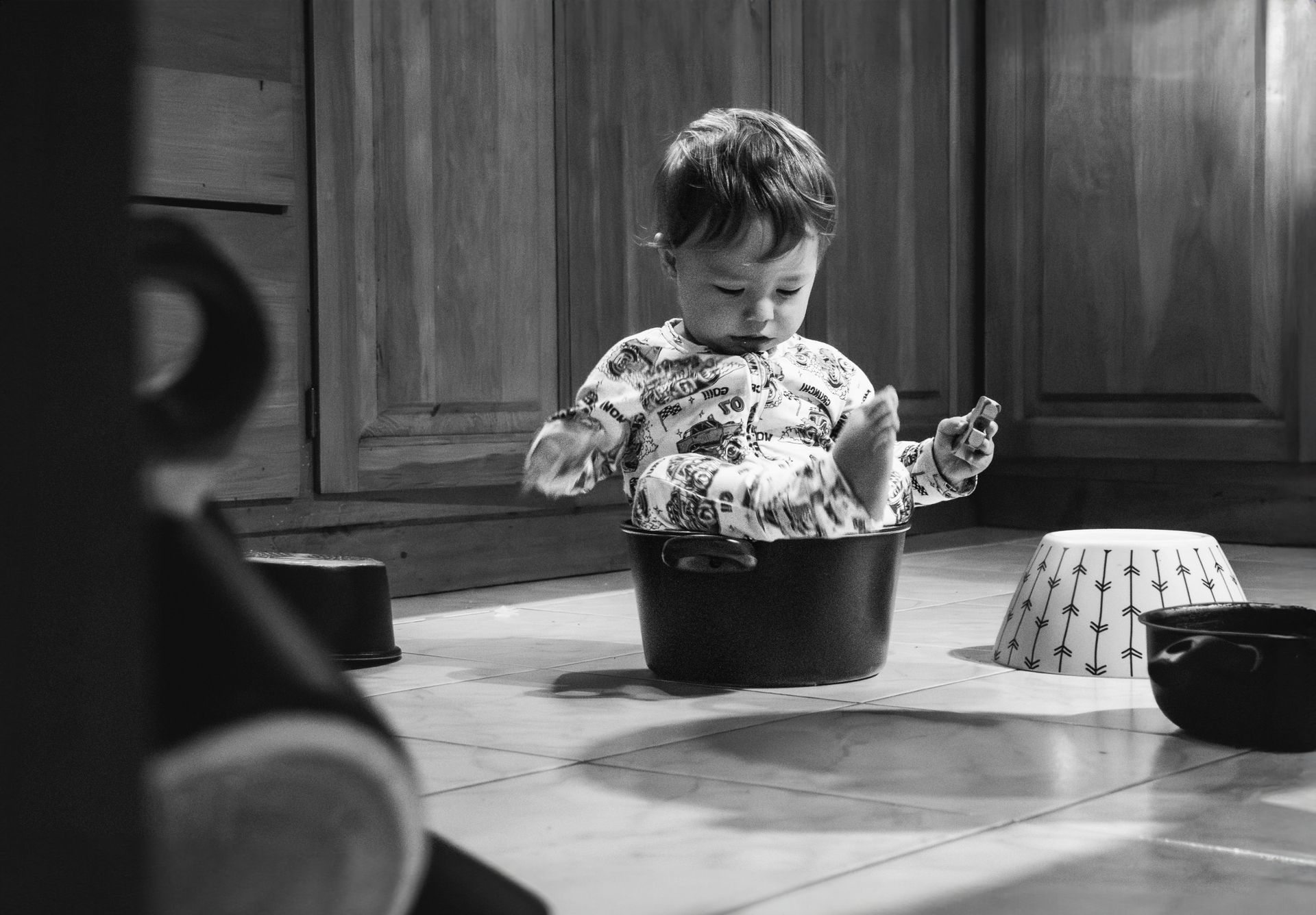 A charming black and white photo of a toddler playing inside a pot in a kitchen setting.