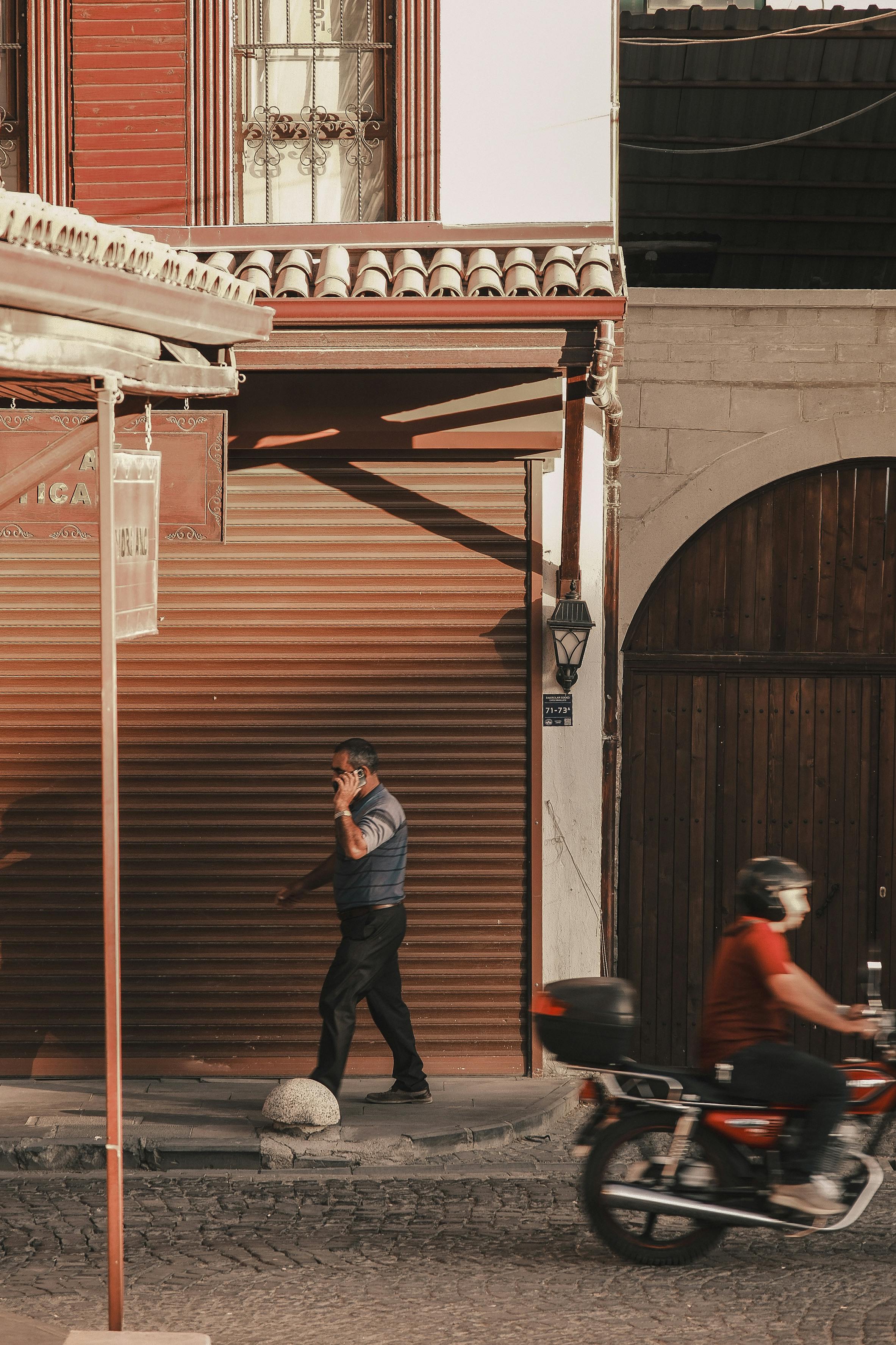 A man talks on the phone on a sunny street with a passing motorcycle, capturing urban life.