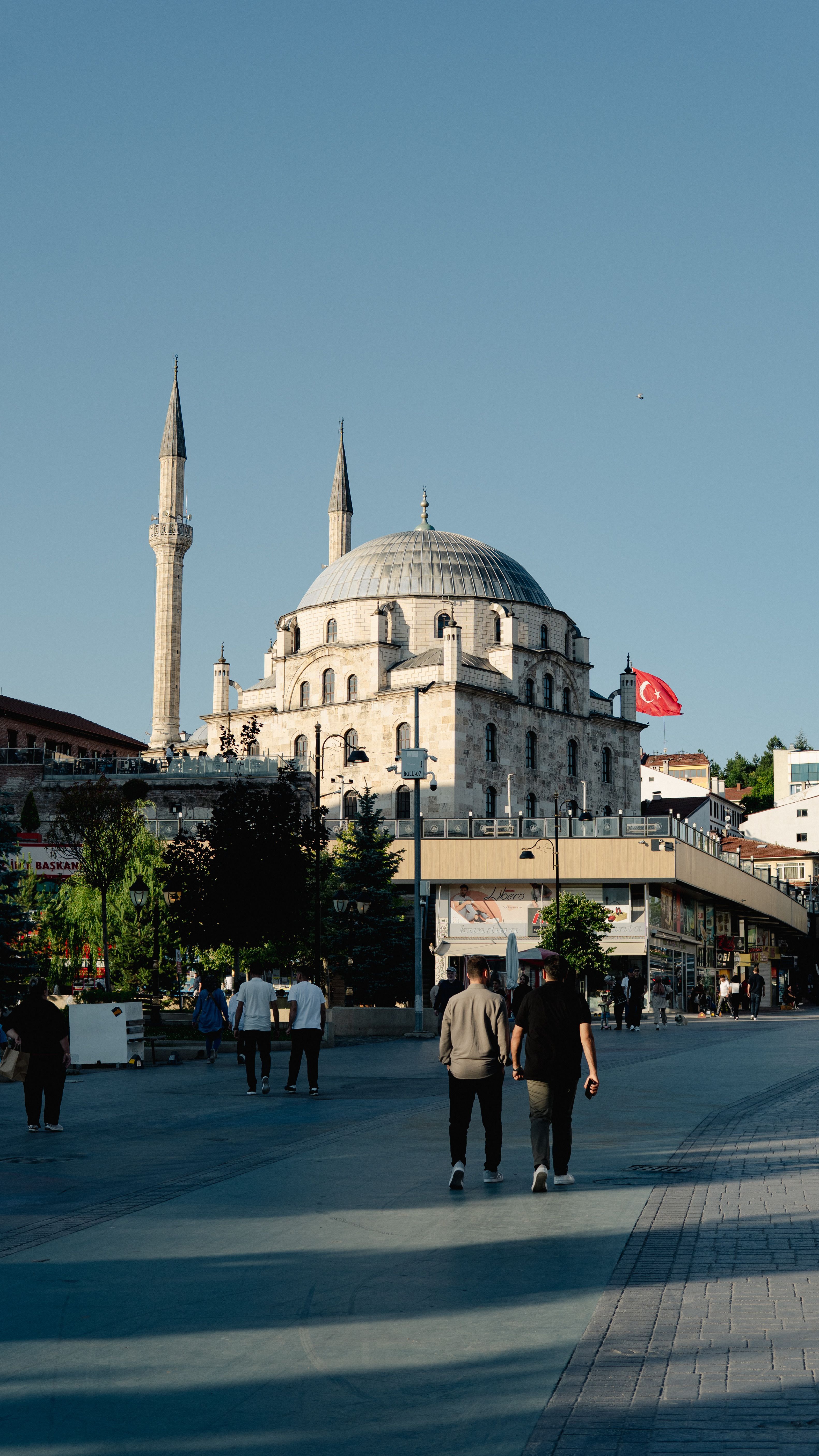 Sunny view of a mosque with minarets and people strolling, showcasing Turkish architecture.