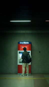 Man using an ATM machine in a dimly lit indoor setting. Night urban scene.
