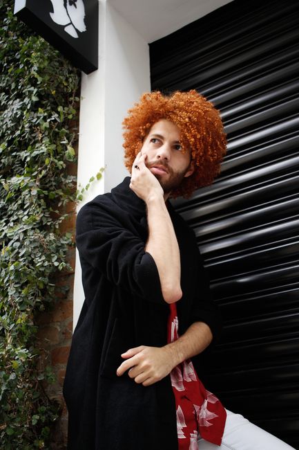 Fashionable man with curly red hair poses thoughtfully outdoors, showcasing unique style and expression.