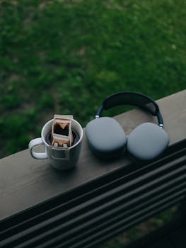 A cup of coffee and headphones rest on a balcony overlooking green grass.