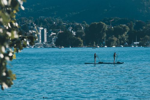 Free Two people paddleboarding on a serene lake with a cityscape and wooded hills in the background. Stock Photo