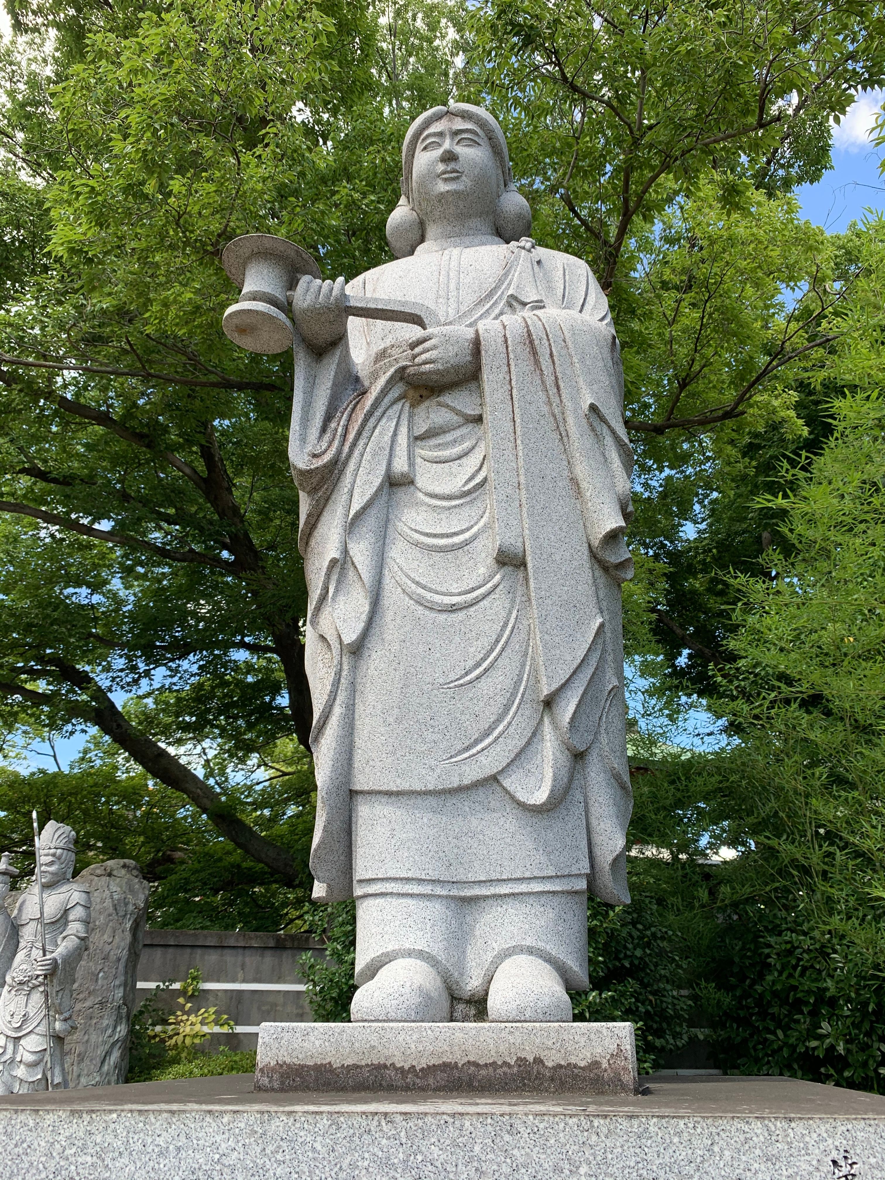 A serene tall stone statue of a Buddhist figure amidst lush green trees on a sunny day.