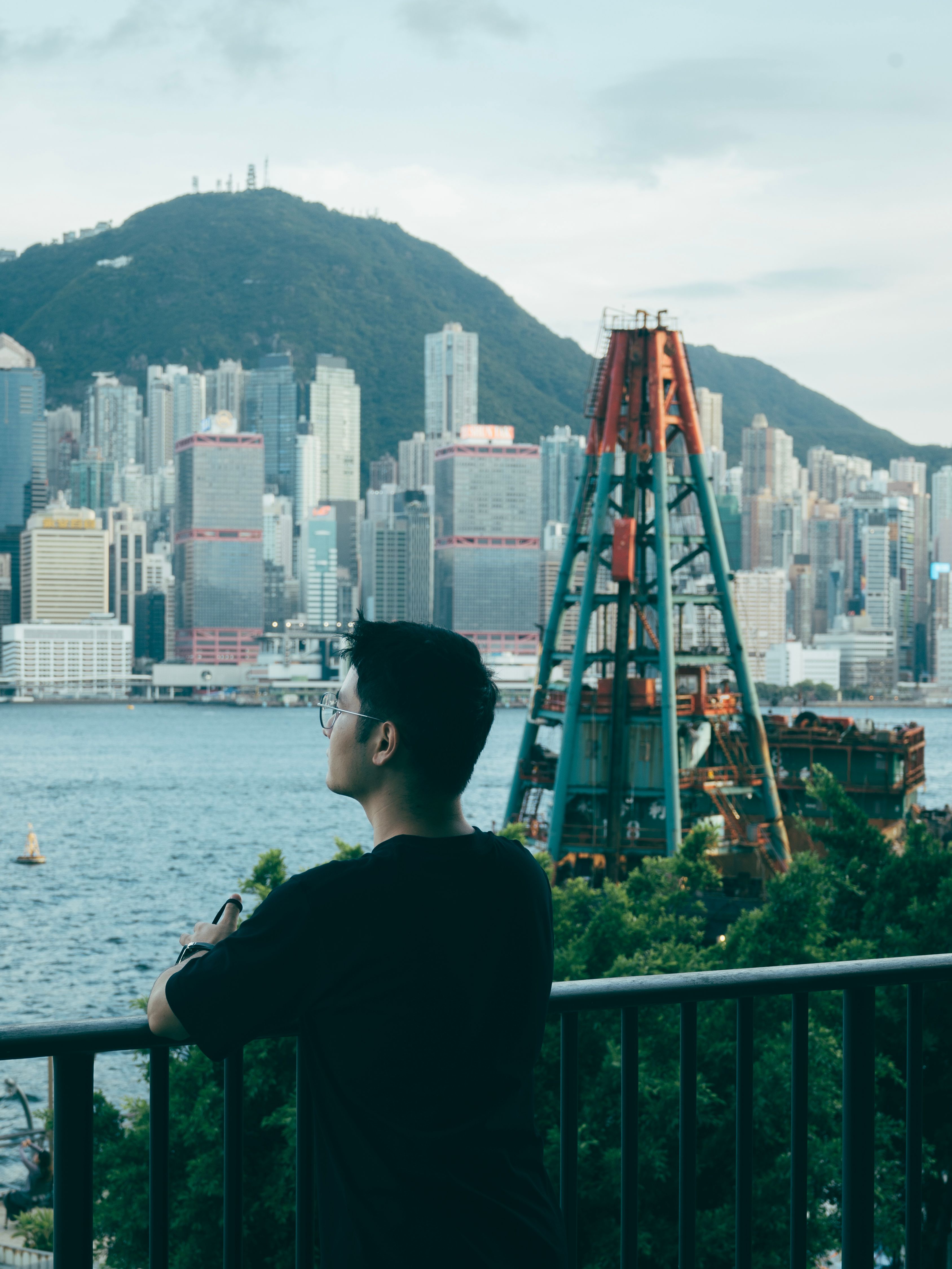 A man leans on a railing gazing at the iconic Hong Kong skyline with Victoria Peak in the background across Victoria Harbour.