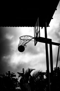Silhouetted basketball hoop with ball in motion against cloudy skies in Pune, India.