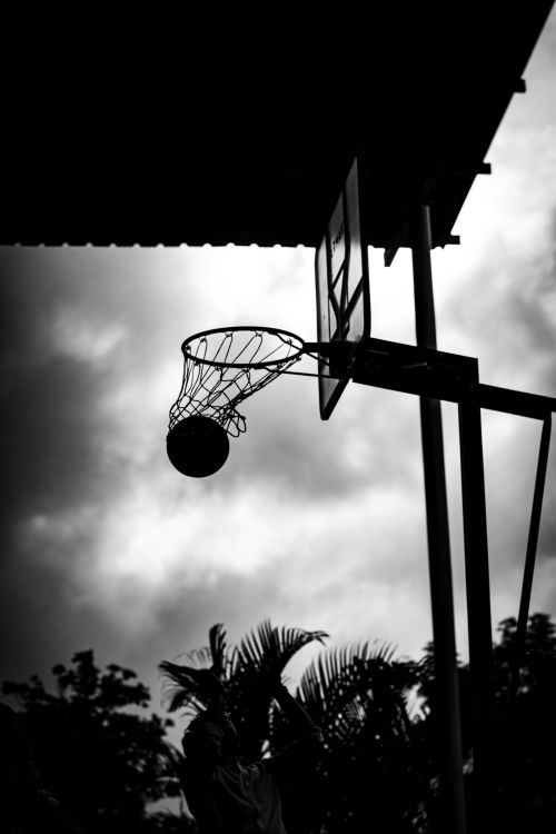 Free Silhouetted basketball hoop with ball in motion against cloudy skies in Pune, India. Stock Photo