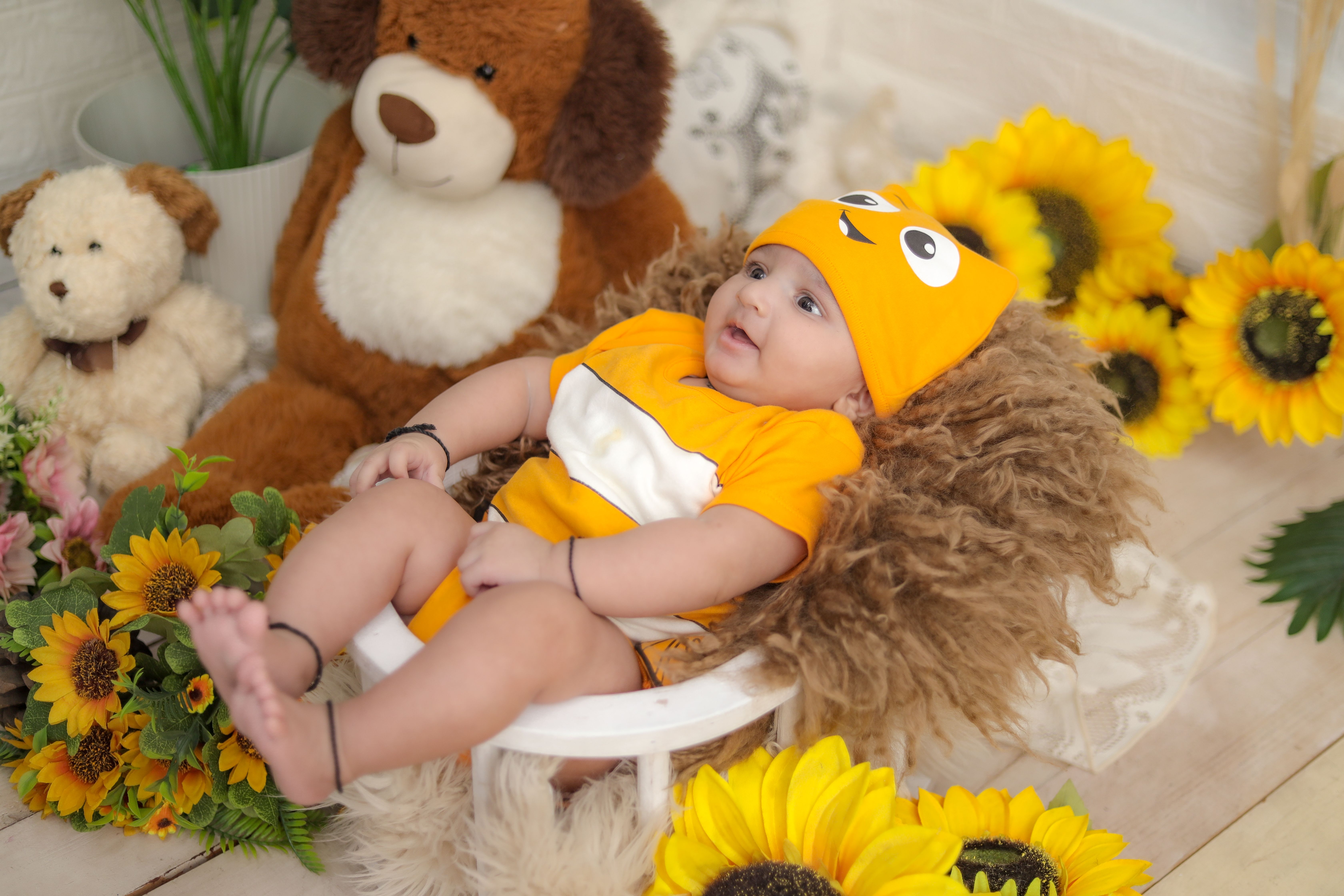 Cute baby dressed in orange outfit surrounded by toys and sunflowers.