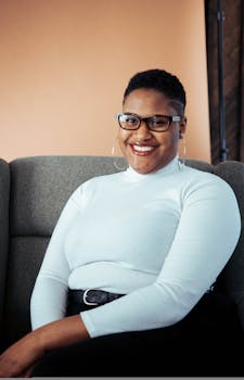 一位时尚的黑人女性戴着眼镜，自信地坐在室内的沙发上微笑