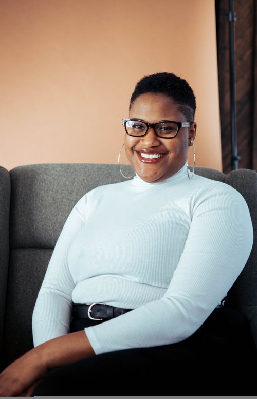 Free A stylish black woman with glasses smiles confidently while sitting on a couch indoors. Stock Photo