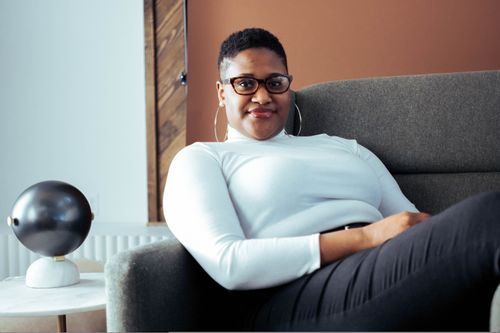 Free Young black woman with glasses sitting on a couch indoors, exuding confidence and style. Stock Photo