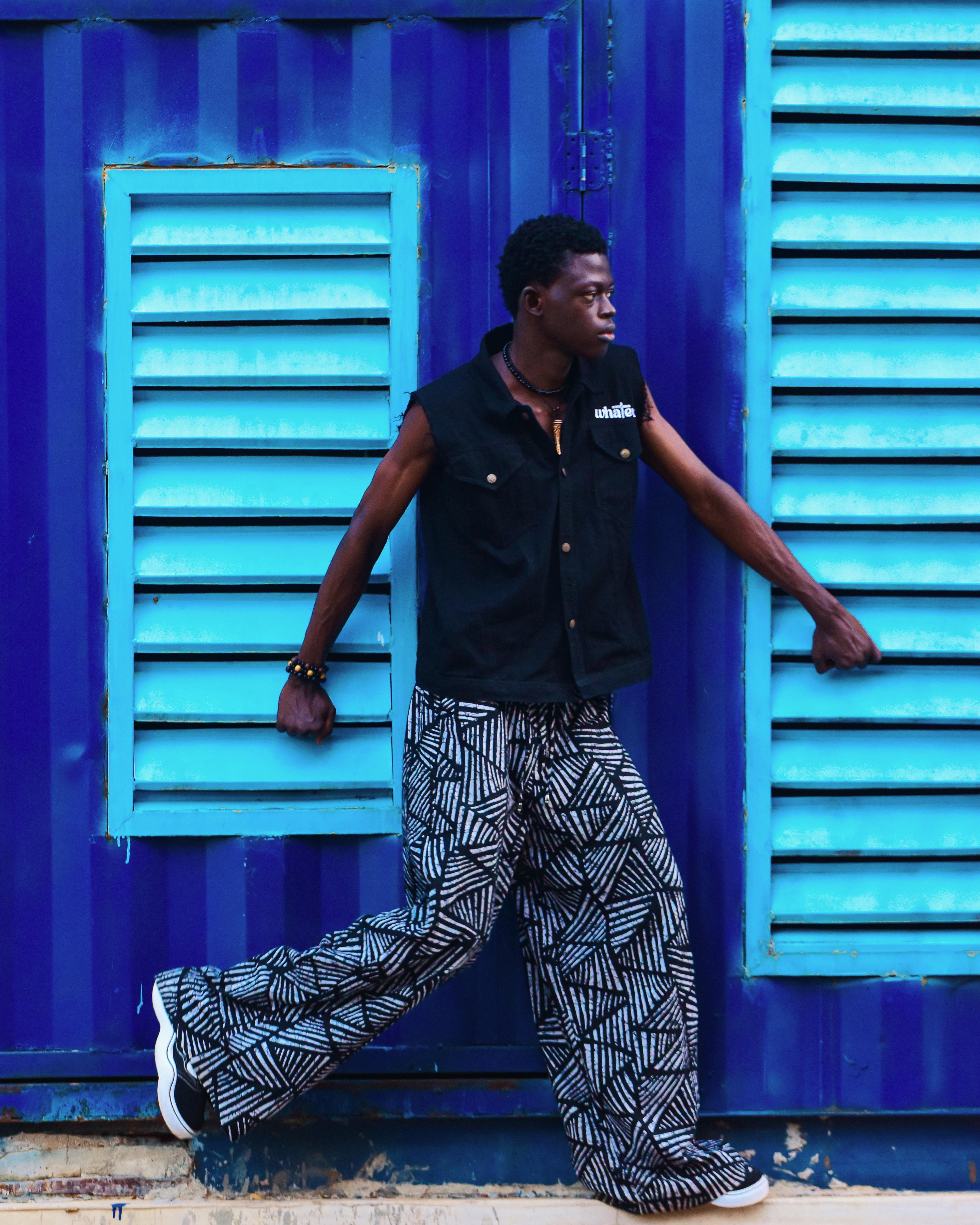 Stylish adult in patterned pants and vest leans against vivid blue shutters.
