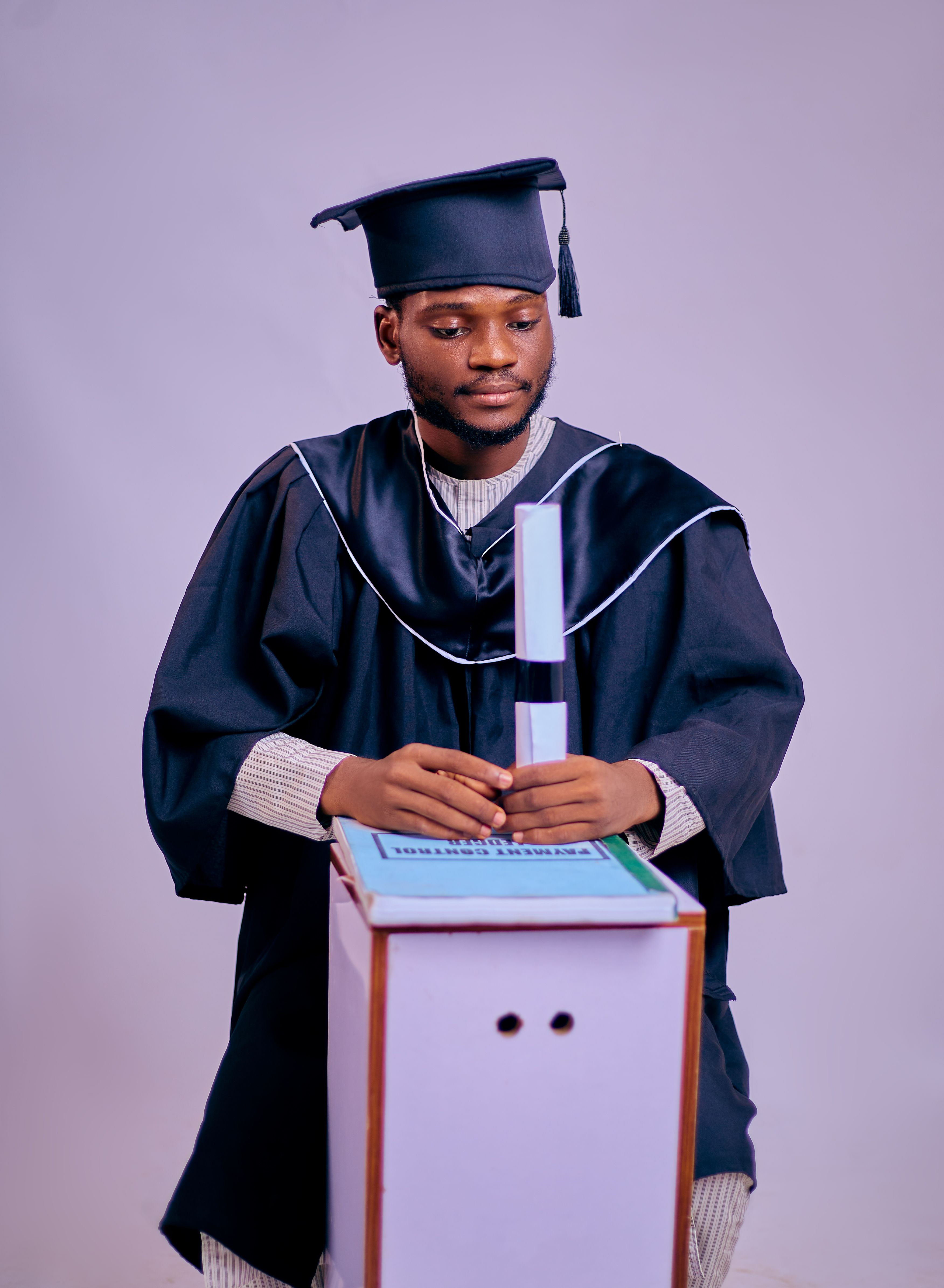 Young adult graduate in cap and gown holding diploma, looking thoughtful.
