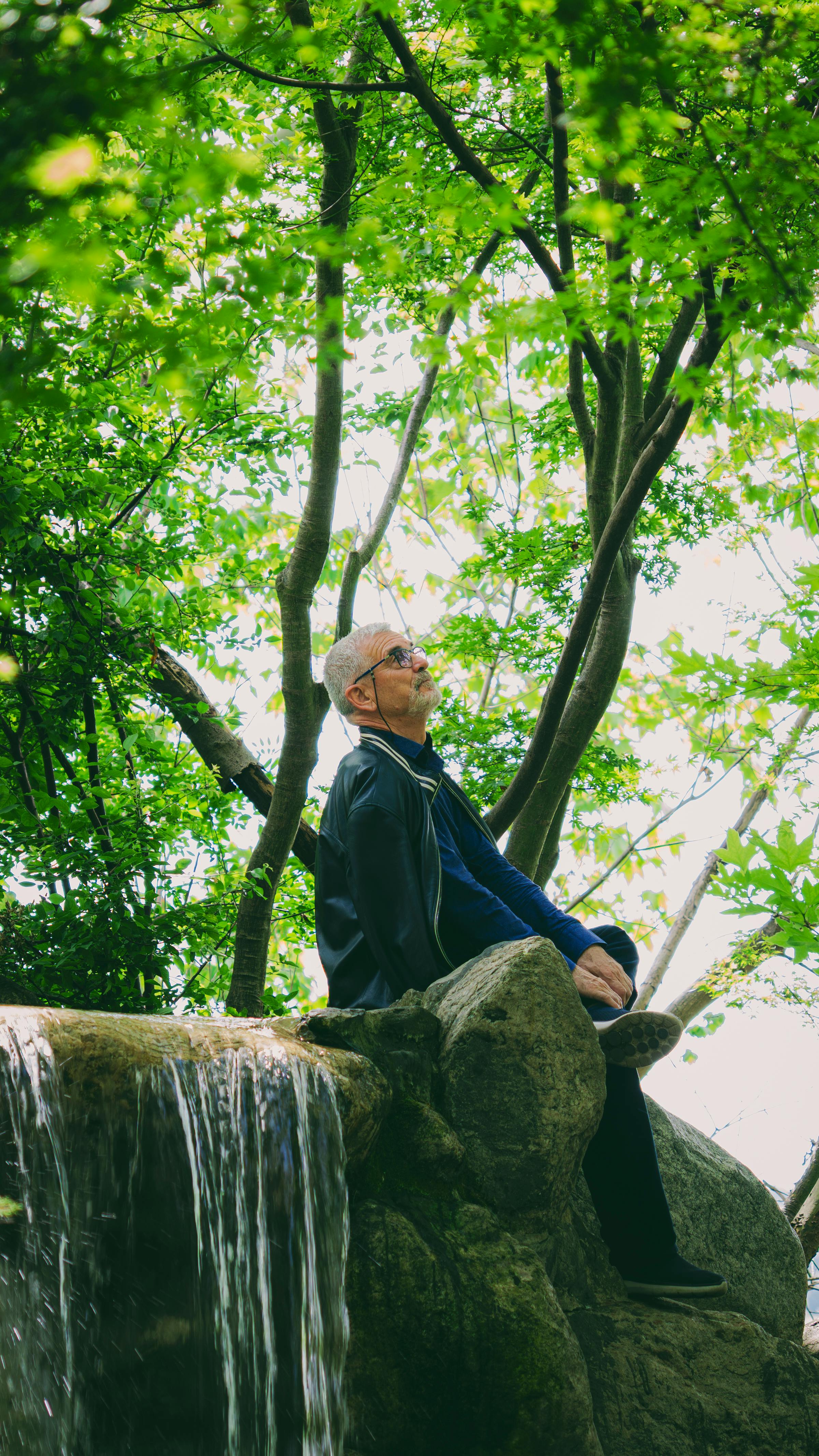 Senior man sitting peacefully by a small waterfall surrounded by lush green trees, enjoying nature's tranquility.