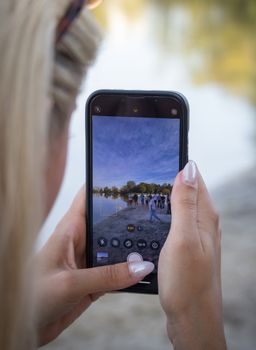 Focus on a smartphone capturing a picturesque lake scene with people in the background, emphasizing photography and technology.