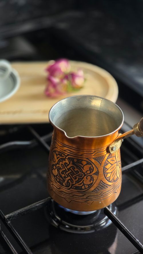 Free Copper Turkish coffee pot with engraved designs heating on a stove, background of pink flowers. Stock Photo