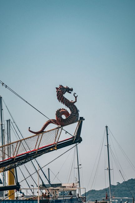 Dragon statue on a ship's prow in Marmaris Harbor with masts and blue sky.