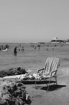 Monochrome beach image featuring a chair, towel, and swimmers in the sea with distant castle.