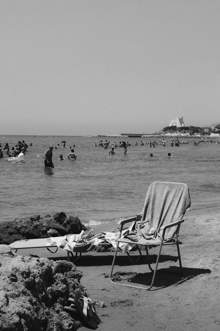 Monochrome beach image featuring a chair, towel, and swimmers in the sea with distant castle.