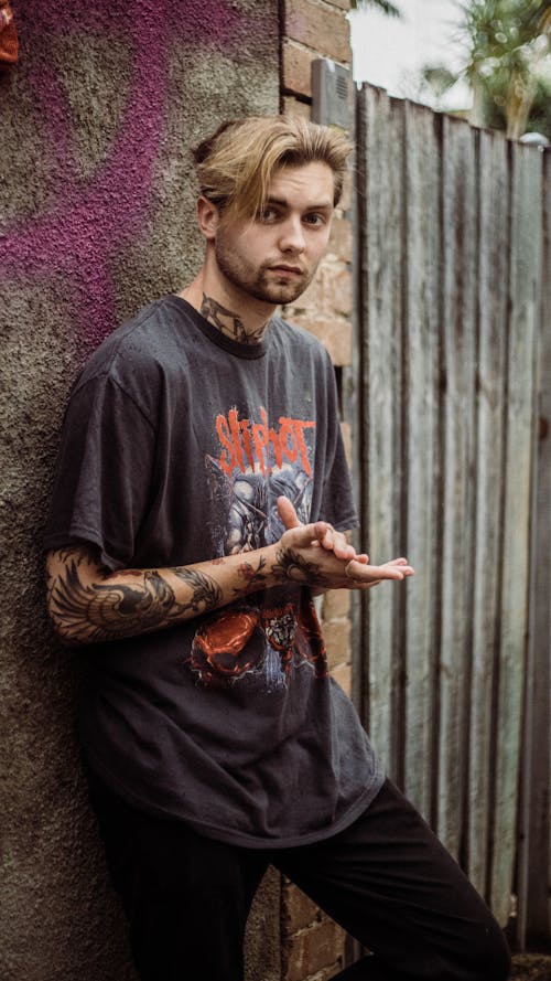 Free Portrait of a stylish, tattooed young man wearing a rock band T-shirt, posing outdoors by a graffiti-covered wall. Stock Photo