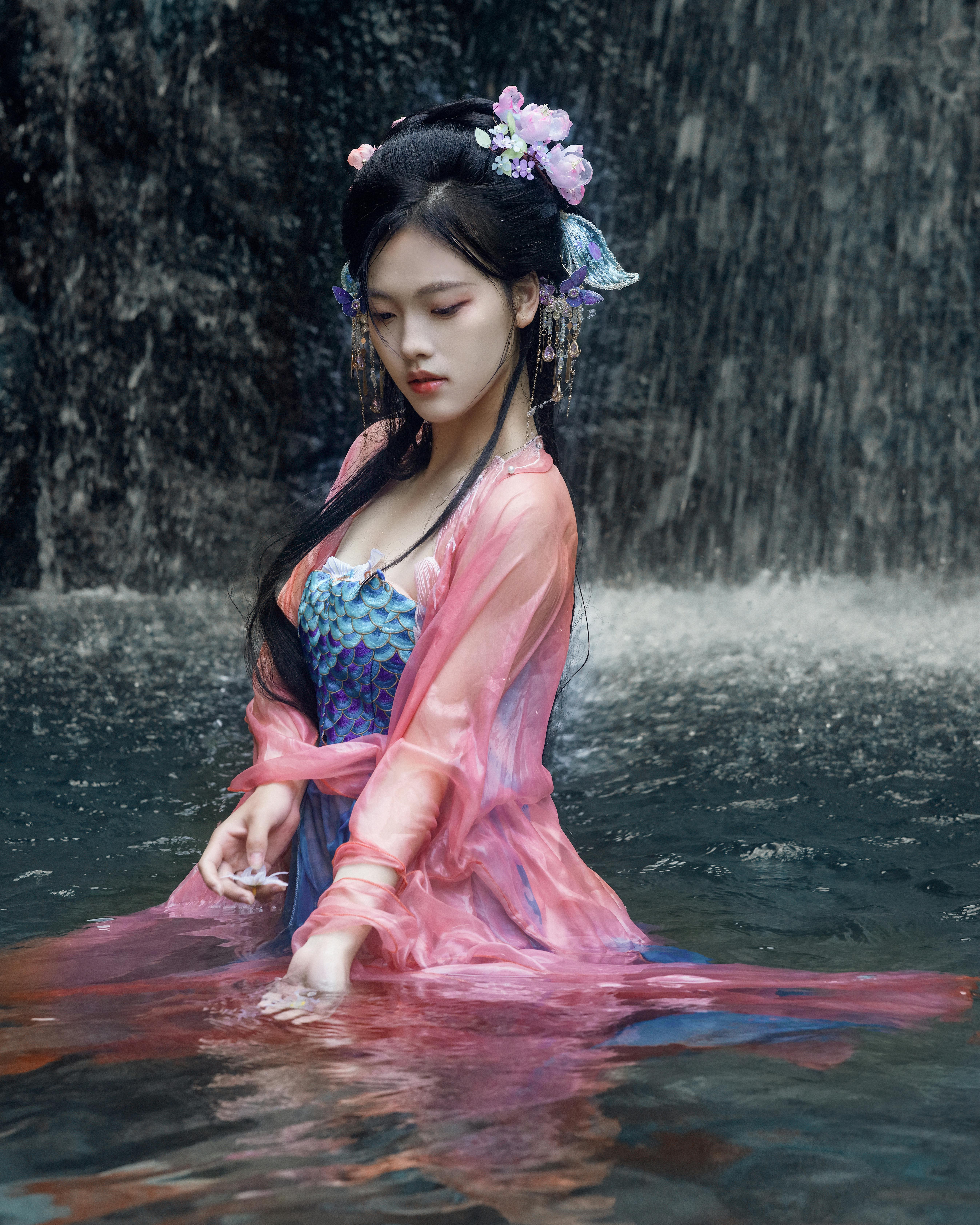A serene woman in colorful traditional attire sits by a cascading waterfall.