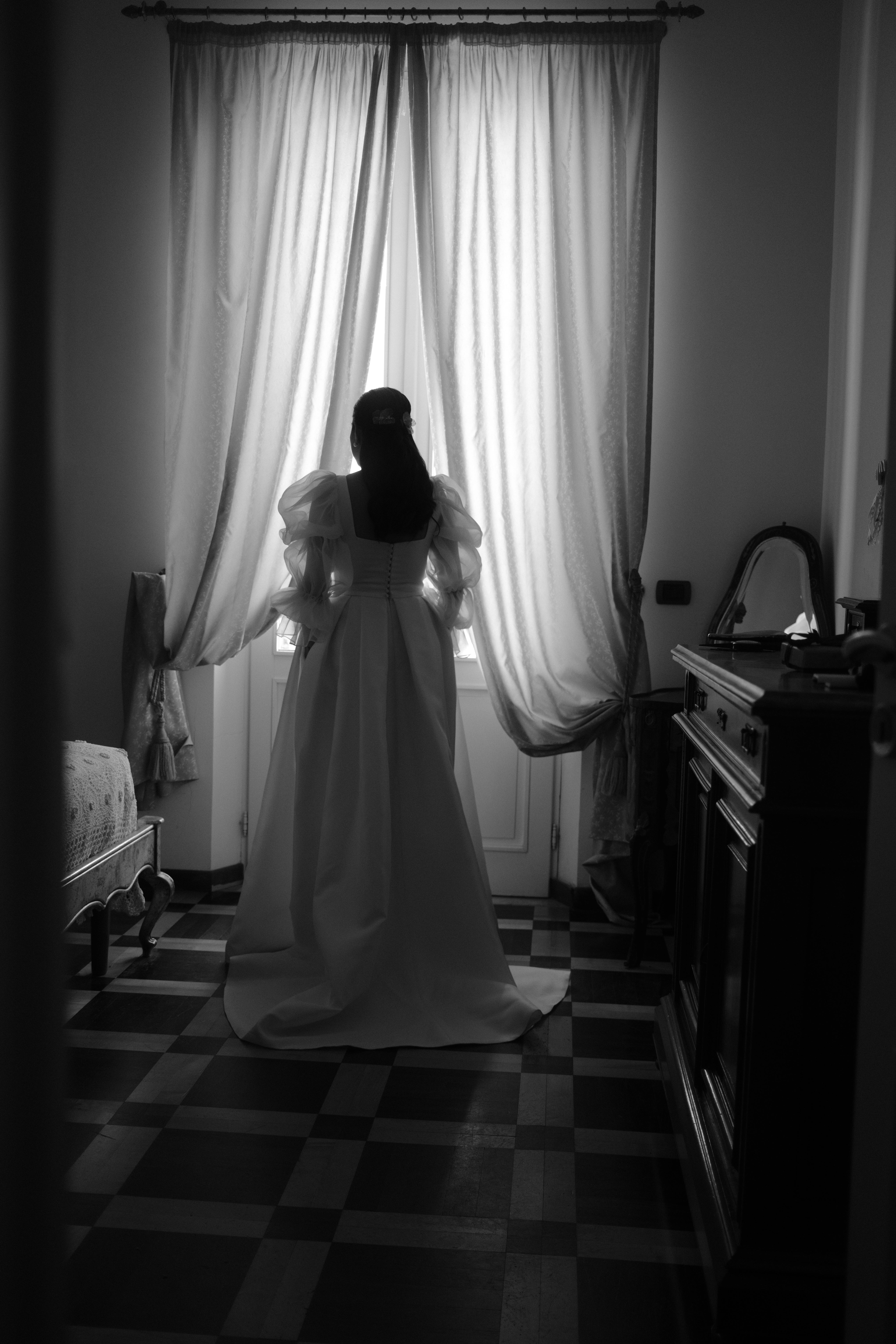 A bride in a classic wedding dress stands in silhouette by a window, exuding elegance.