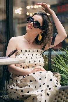 Stylish woman wearing sunglasses and a polka dot dress sitting at an outdoor cafe table.