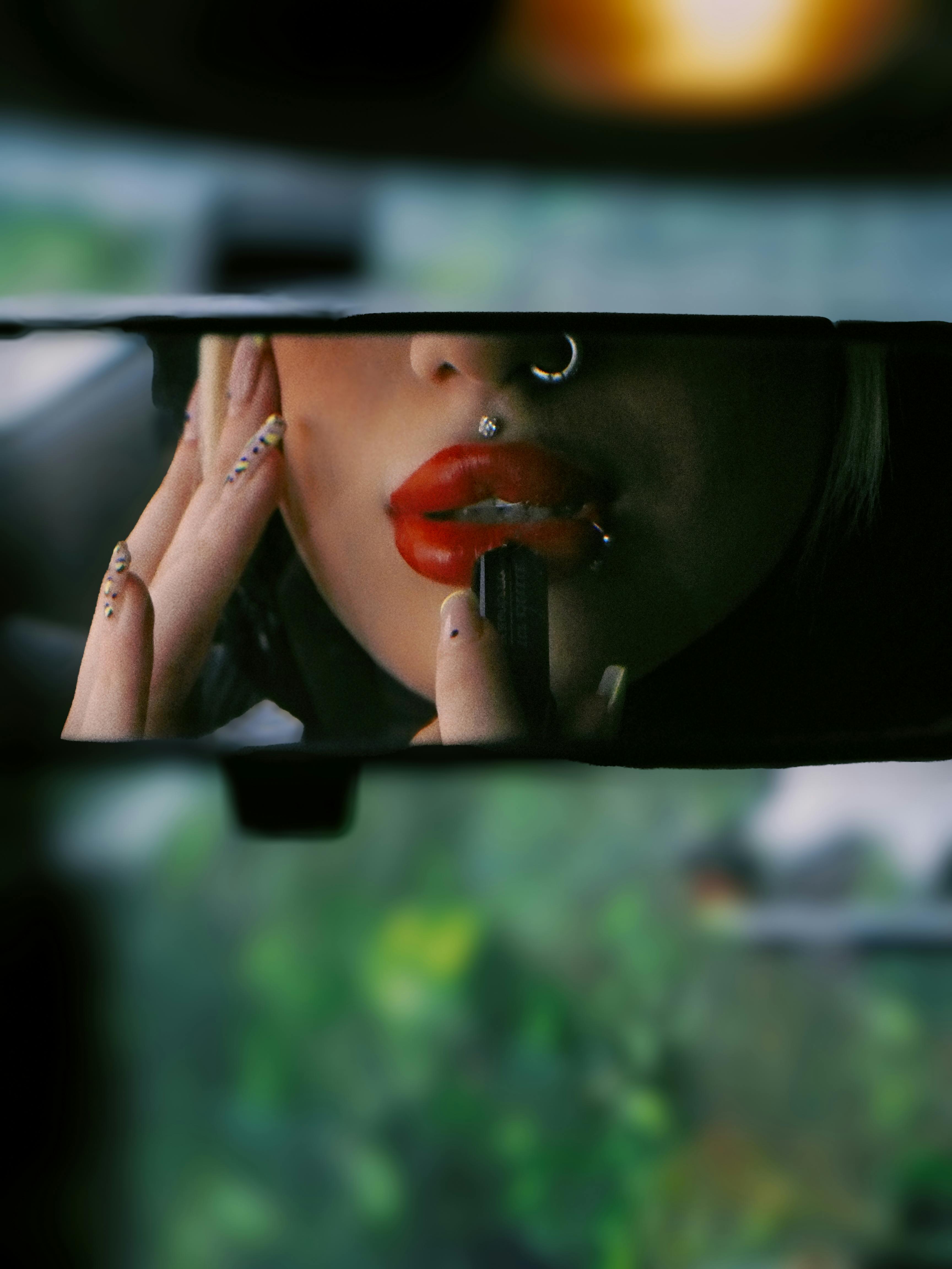 Woman applying lipstick in car rearview mirror, focusing on vibrant lips and stylish accessories.