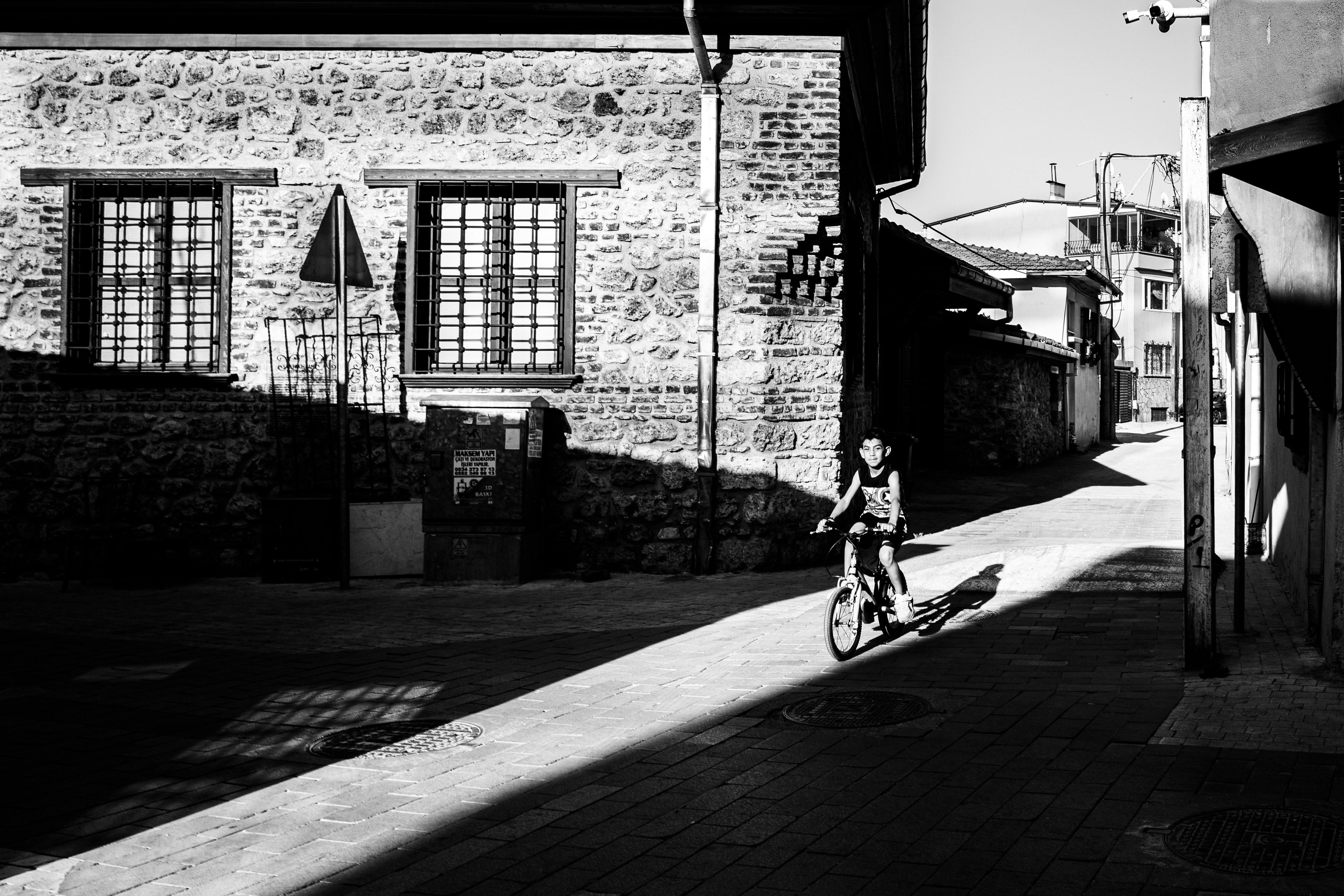 Black and white image depicting a child riding a bicycle through a shadowy street in Bursa, Türkiye.