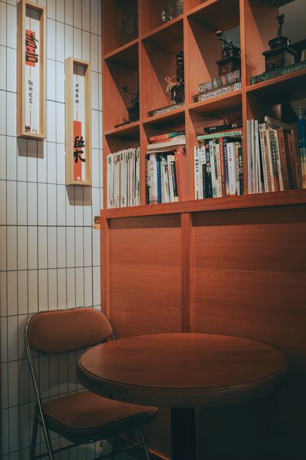 A warmly lit, cozy indoor corner with a round table, chair, and wooden shelves filled with books.