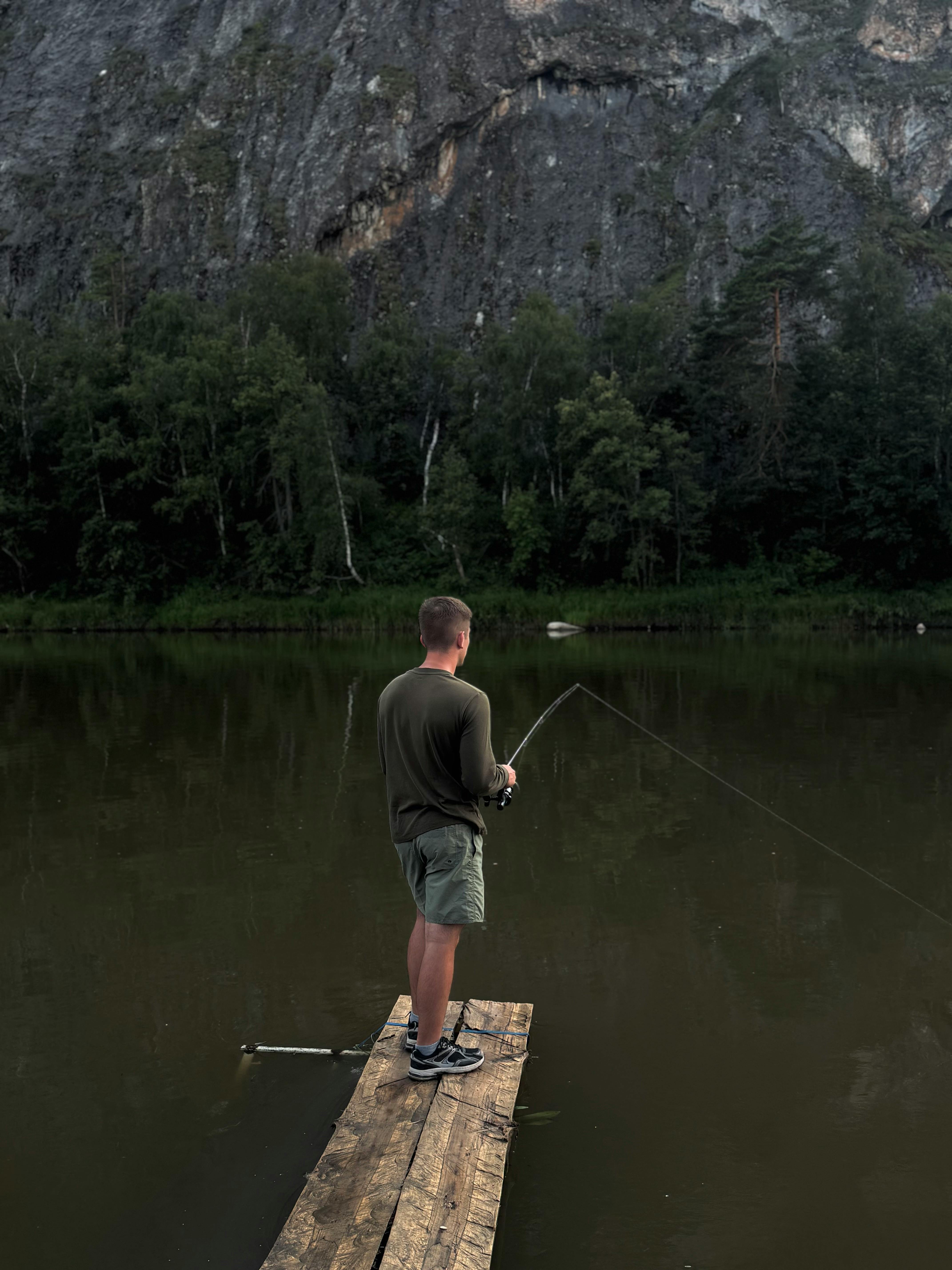 Adult man fishing from a wooden dock by a lake with a forest and rocky cliff in the background.